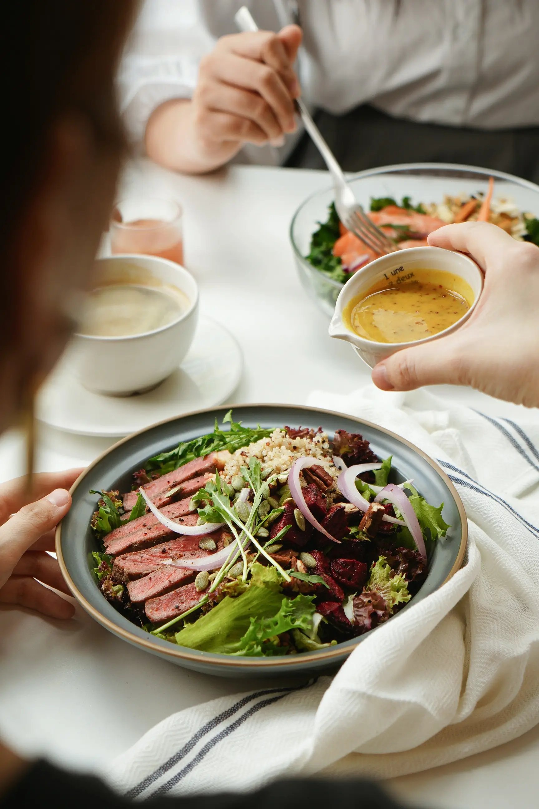 close-up of a fresh salad bowl topped with sliced steak, mixed greens, quinoa, beetroot, red onion, seeds, and nuts, while someone pours mustard dressing over it; another person eats a similar salad in the background, with coffee and juice on the table.