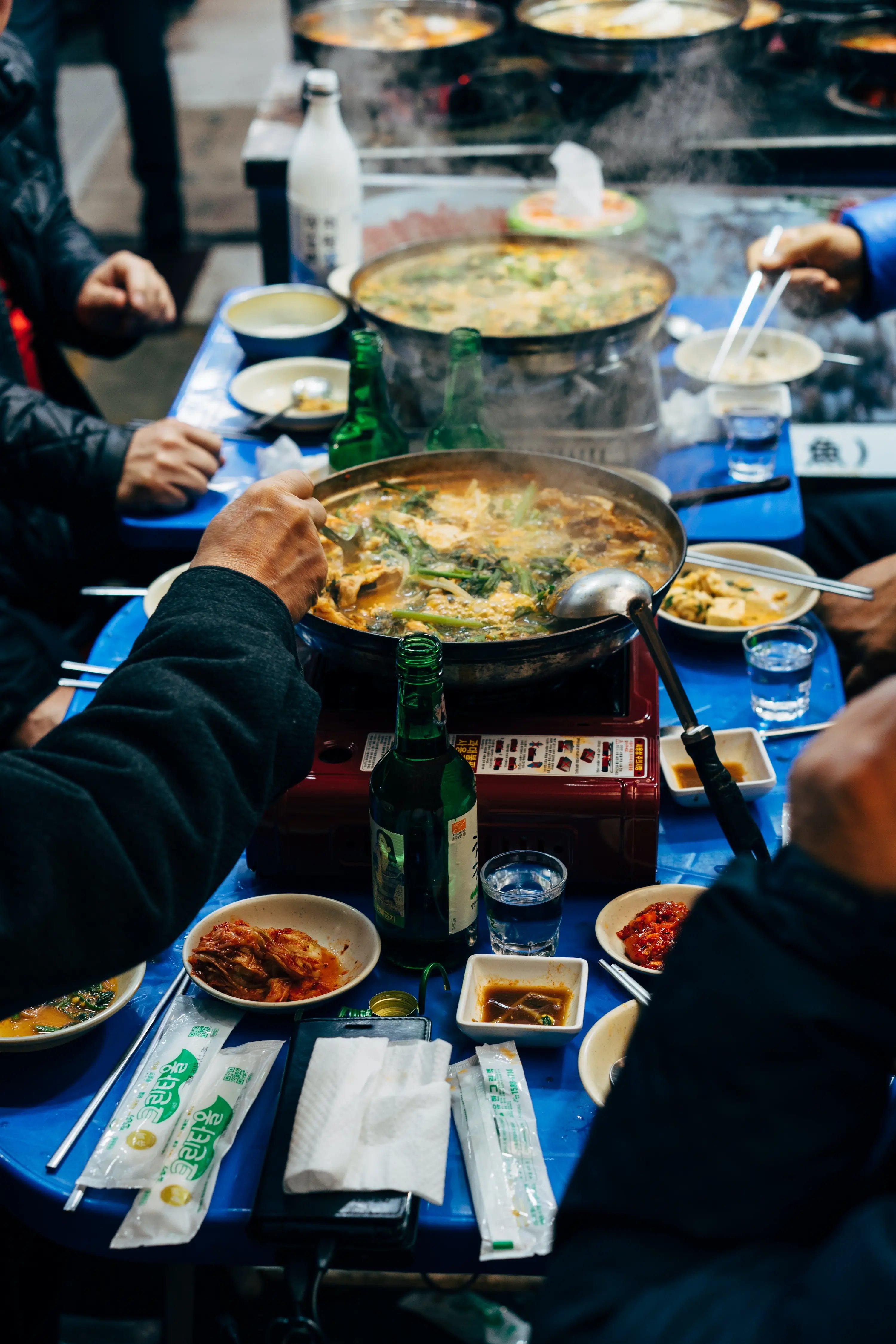 street-food scene with several people gathered around a small blue plastic table, cooking a large bubbling pot of stew on a portable gas burner; side dishes like kimchi and sauces surround the pot, along with bottles of soju, bowls, chopsticks, and napkins, capturing a busy, casual outdoor dining atmosphere.
